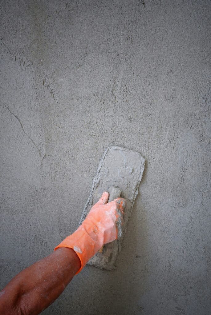 Enduit ciment et maçonnerie : réussir l'application sur mur. Maçon appliquant du ciment sur un mur brut avec une truelle. Gros plan sur la main gantée orange et le mortier gris.